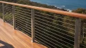 Modern residential timber deck with a premium, compliant stainless steel wire rope balustrade featuring 11 horizontal cable runs, vertical metal posts, and a continuous timber top rail overlooking a coastal landscape view.